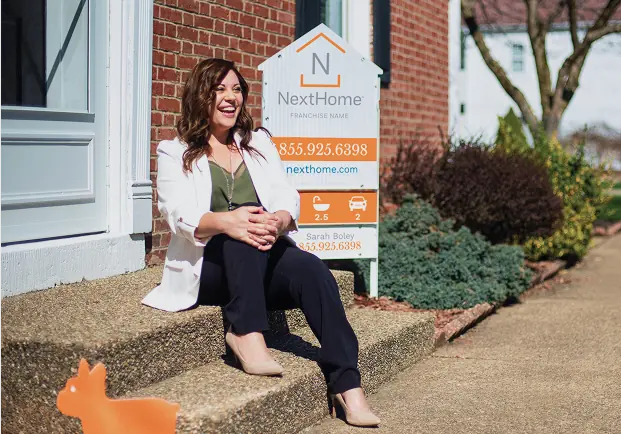 Person sitting on home steps in front of a home sale sign.